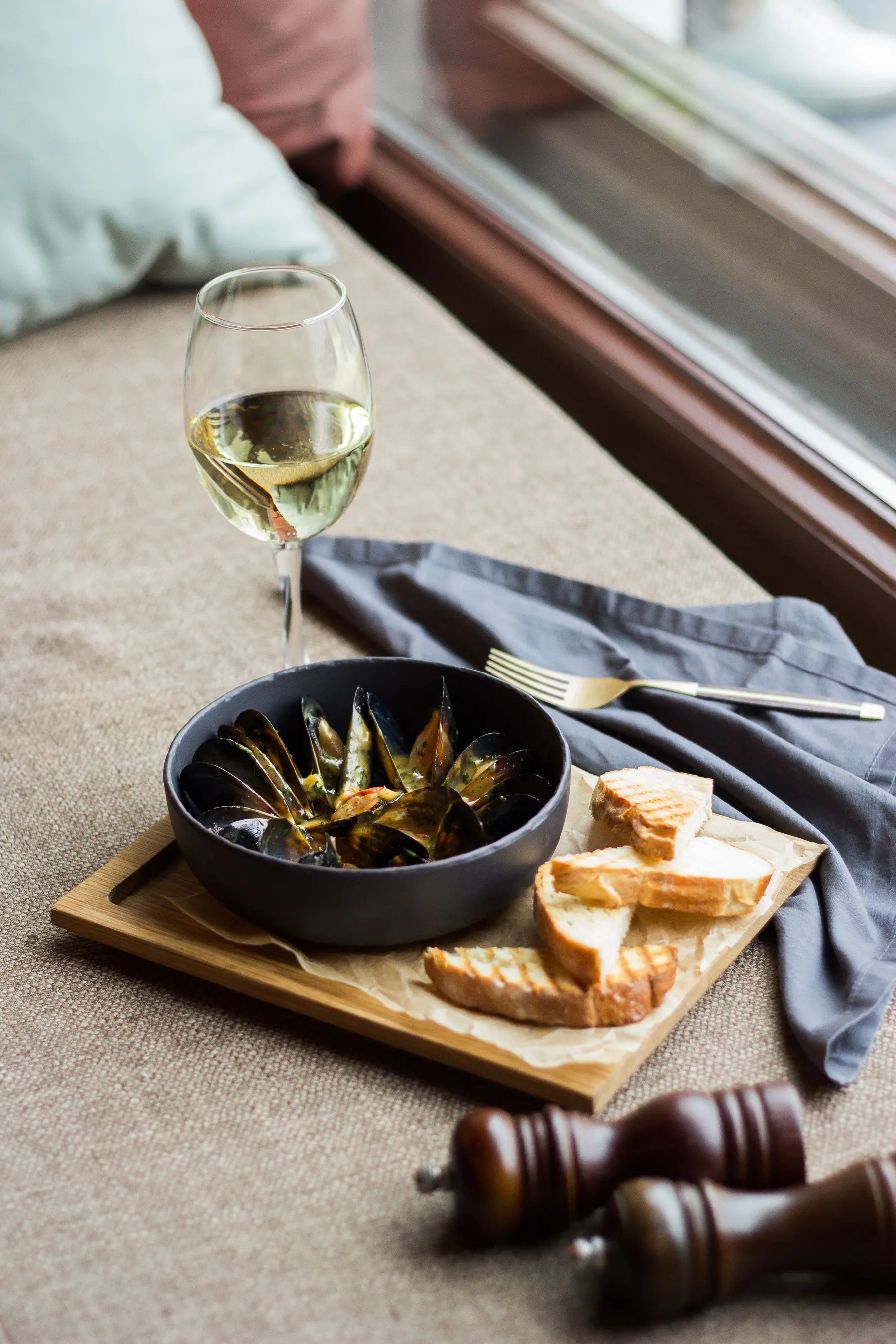 A glass of white wine with olives and bread by a window